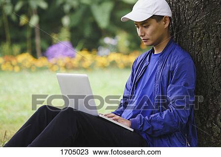 Young man using laptop while sitting by tree at park View Large Photo Image Stock Image - Young man using laptop while sitting by tree at park. Fotosearch