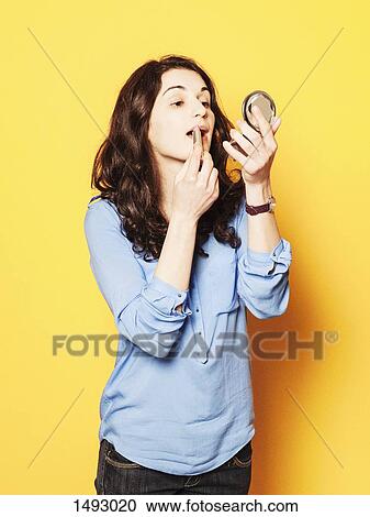Stock Image - Young woman applying lipstick against yellow background. Fotosearch