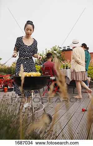 Young woman barbecuing corns while standing on patio with friends in background View Large Photo Image Picture - Young woman barbecuing corns while standing on patio with friends in background. Fotosearch