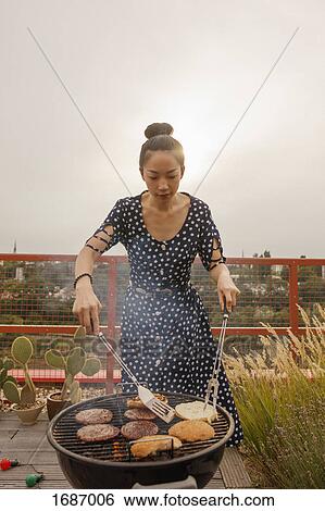 Stock Photograph - Young woman barbecuing steak and buns at patio against clear sky. Fotosearch