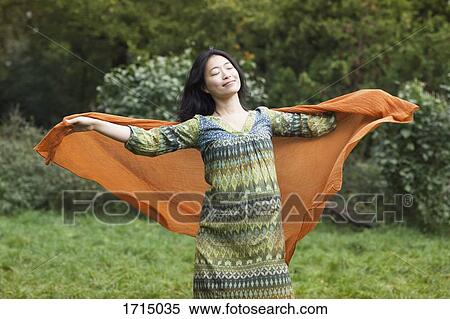 Young woman holding orange scarf while standing with arms outstretched against trees at park View Large Photo Image Stock Photography - Young woman holding orange scarf while standing with arms outstretched against trees at park. Fotosearch