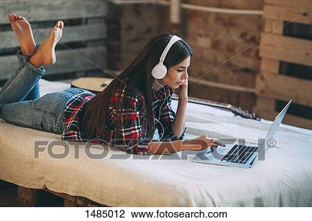 Young woman listening music while using laptop on bed at home View Large Photo Image Stock Image - Young woman listening music while using laptop on bed at home. Fotosearch