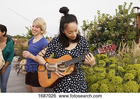 Stock Photograph - Young woman playing guitar on patio while female friends standing in background. Fotosearch