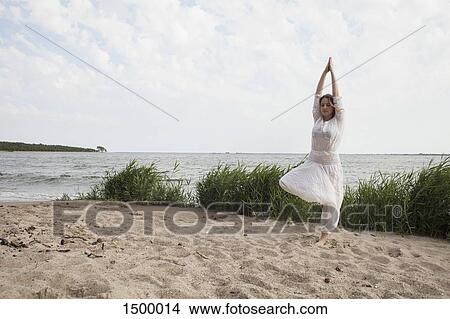 Picture - Young woman practicing yoga on sea shore against sky. Fotosearch