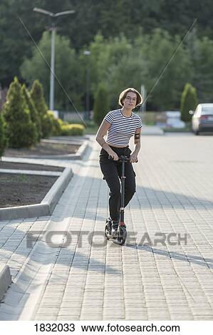 Stock Image - Young woman riding push scooter on sunny road. Fotosearch