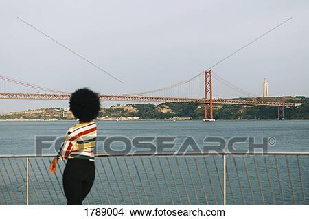 Young woman standing at waterfront, looking at 25 de Abril Bridge and Cristo Rei Statue, Lisbon View Large Photo Image Picture - Young woman standing at waterfront, looking at 25 de Abril Bridge and Cristo Rei Statue, Lisbon. Fotosearch