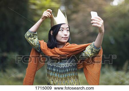 Young woman wearing crown taking selfie through mobile phone while standing at park View Large Photo Image Stock Photo - Young woman wearing crown taking selfie through mobile phone while standing at park. Fotosearch