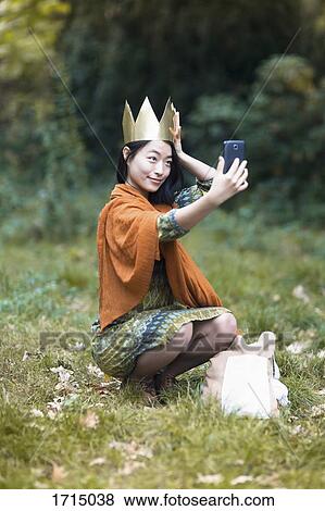 Young woman wearing crown taking selfie through mobile phone while crouching on field at park View Large Photo Image Stock Photo - Young woman wearing crown taking selfie through mobile phone while crouching on field at park. Fotosearch