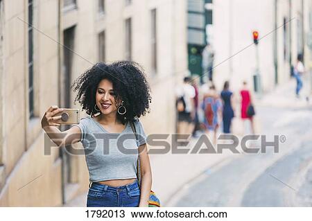 Stock Image - Young woman with smart phone taking selfie on urban street. Fotosearch