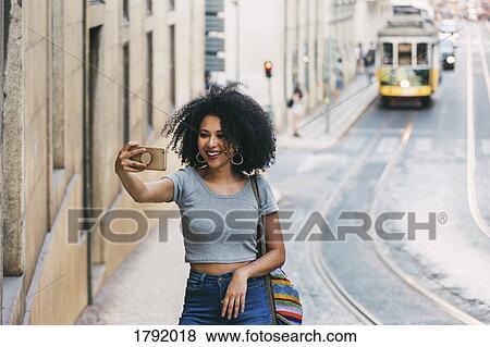 Stock Photo - Young woman with smart phone taking selfie on urban street. Fotosearch