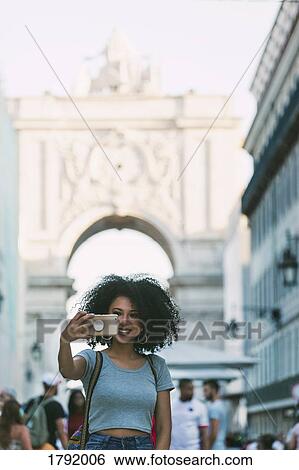 Stock Photograph - Young woman with smart phone taking selfie with Rua Augusta Arch, Lisbon, Portugal. Fotosearch