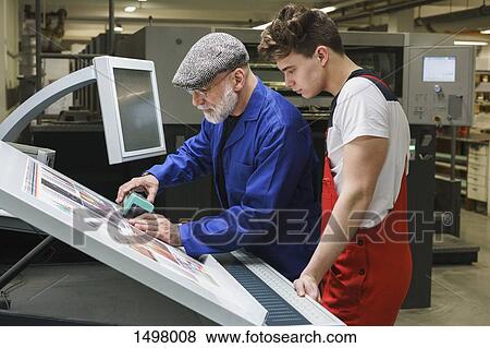 Stock Photo - Young worker looking at mature colleague doing quality check of printout with scanner. Fotosearch