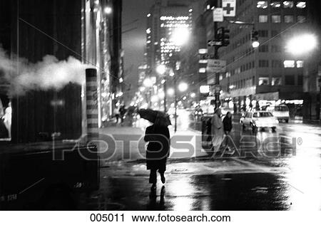 Stock Image - A person crossing a New York city street with an umbrella at night. Fotosearch