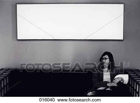 Stock Image - a woman sitting on a couch with white painting behind her. Fotosearch