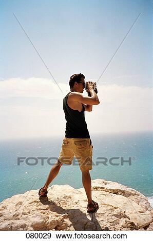Stock Photo - A man on top of a cliff looking through a camera, Australia. Fotosearch