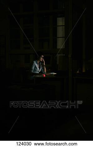 Stock Image - Woman sitting at office desk in the dark. Fotosearch