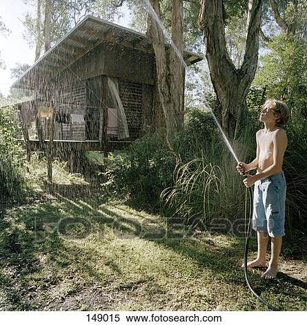 Stock Photography - A boy spraying water near a forest hut. Fotosearch