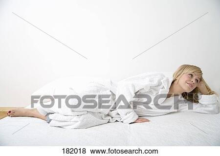 Stock Photo - Young woman in bathrobe looking away while lying in bed on her side. Fotosearch
