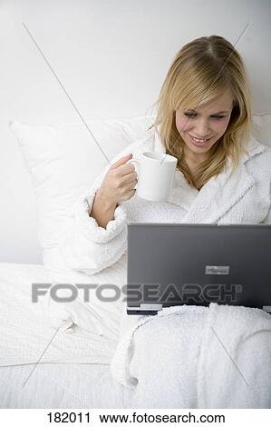 Young woman in bathrobe sitting in bed with laptop and coffee cup View Large Photo Image Stock Image - Young woman in bathrobe sitting in bed with laptop and coffee cup. Fotosearch