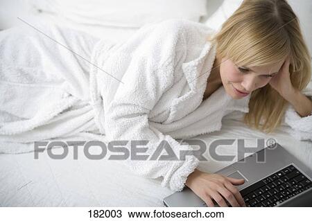 Young woman lying down on a bed in a bathrobe and using a laptop View Large Photo Image Stock Image - Young woman lying down on a bed in a bathrobe and using a laptop. Fotosearch