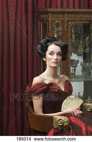 Picture - Elegant young woman seated in red ball gown and holding fan. Fotosearch