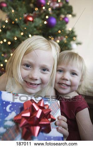 Stock Image - Two young girls holding present near Christmas tree. Fotosearch