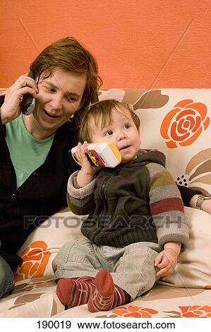 Mother and baby boy both sitting on sofa using a phone View Large Photo Image Stock Photo - Mother and baby boy both sitting on sofa using a phone. Fotosearch