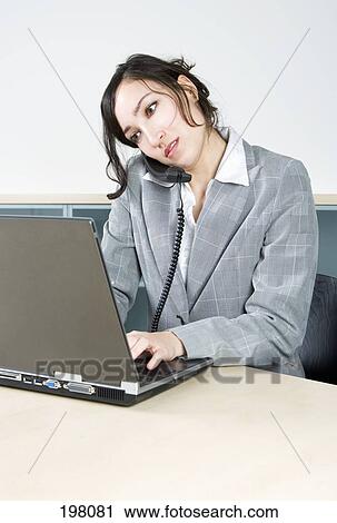 Stock Image - A woman working on a laptop while talking on the telephone. Fotosearch