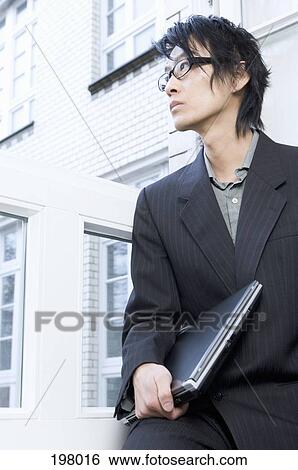 An Asian businessman holding a laptop next to a window View Large Photo Image Stock Photograph - An Asian businessman holding a laptop next to a window. Fotosearch