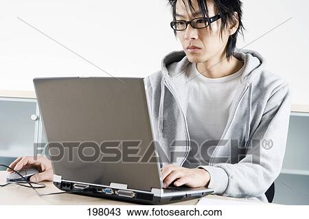 Stock Image - An Asian man working on a laptop in an office setting. Fotosearch