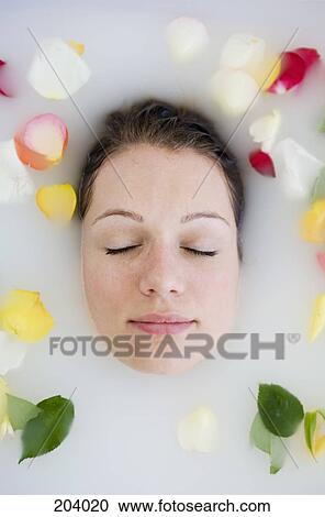 Stock Image - Young woman with eyes closed lying in bathtub and surrounded by rose petals. Fotosearch