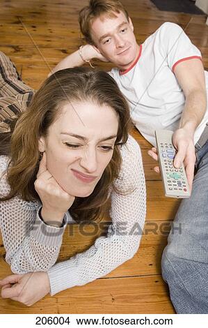 Young woman making face whilst lying down next to young man holding remote control View Large Photo Image Picture - Young woman making face whilst lying down next to young man holding remote control. Fotosearch