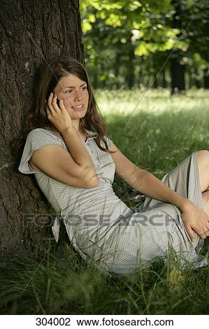 Stock Image - Girl sitting below tree, talking on mobile phone. Fotosearch