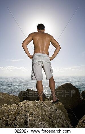 Stock Photo - Rear view of man standing on rock in front of sea. Fotosearch
