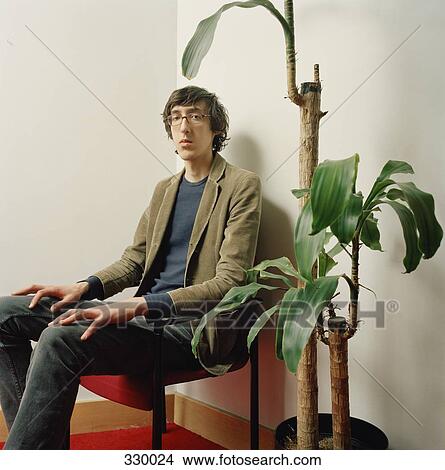 Picture - Young man sitting on chair next to potted plant. Fotosearch