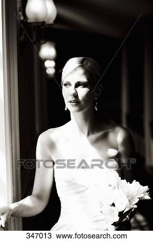 Stock Image - Bride holding wedding bouquet and looking out window. Fotosearch