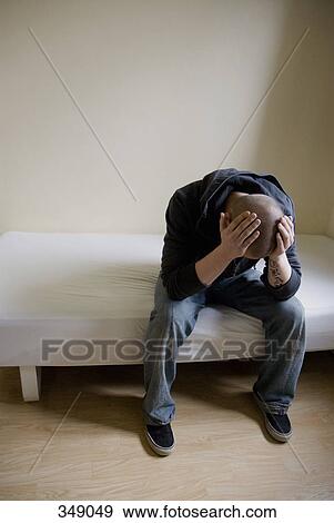 Stock Photo - Young man sitting on bed holding his head in his hands. Fotosearch
