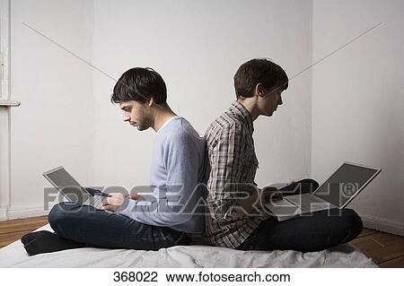 Stock Image - Two young men sitting back to back using laptops. Fotosearch