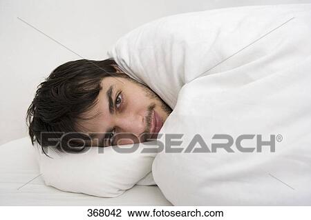 Stock Image - Young man lying down wrapped in duvet. Fotosearch