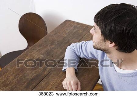 Stock Image - Young man sitting at table looking at empty chair. Fotosearch
