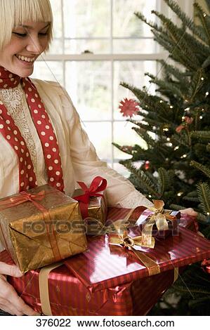 Stock Image - Young woman holding presents and standing by a Christmas tree. Fotosearch
