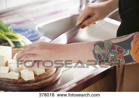 Stock Photo - Woman chopping tofu in kitchen with ornate tattooed arm. Fotosearch