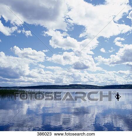 Stock Image - Man standing in a lake surrounded by mountains. Fotosearch