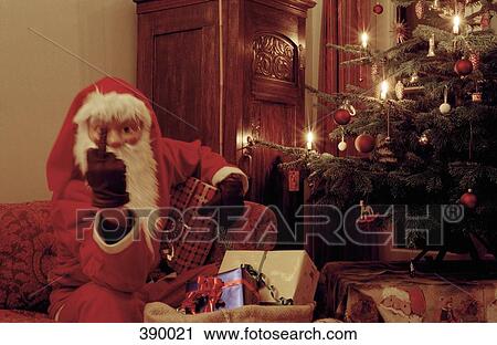 Stock Image - Santa Claus raising his middle finger whilst sitting on a sofa next to an illuminated Christmas tree. Fotosearch