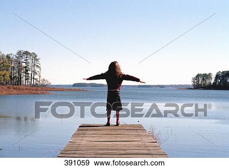 Stock Photo - Young girl standing at the edge of a wooden jetty with her arms outstretched. Fotosearch