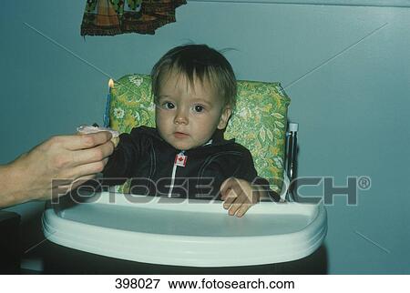 Stock Photo - Young boy sitting in a highchair in front of a cupcake with a lit candle. Fotosearch