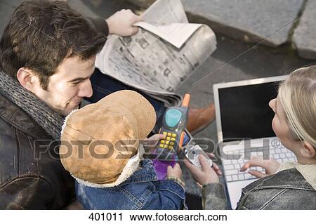Stock Image - Family sitting outside using laptop and mobile phone. Fotosearch
