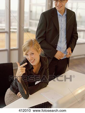 A young woman sitting at an office desk and pointing as a young man stands beside her View Large Photo Image Stock Image - A young woman sitting at an office desk and pointing as a young man stands beside her. Fotosearch