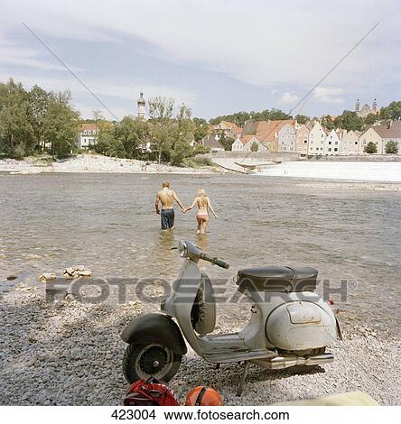 Picture - A motor scooter parked on a river bank whilst a young couple go into the water. Fotosearch