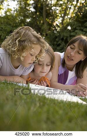 Stock Image - A woman and two girls lying down in a backyard and reading a book together. Fotosearch
